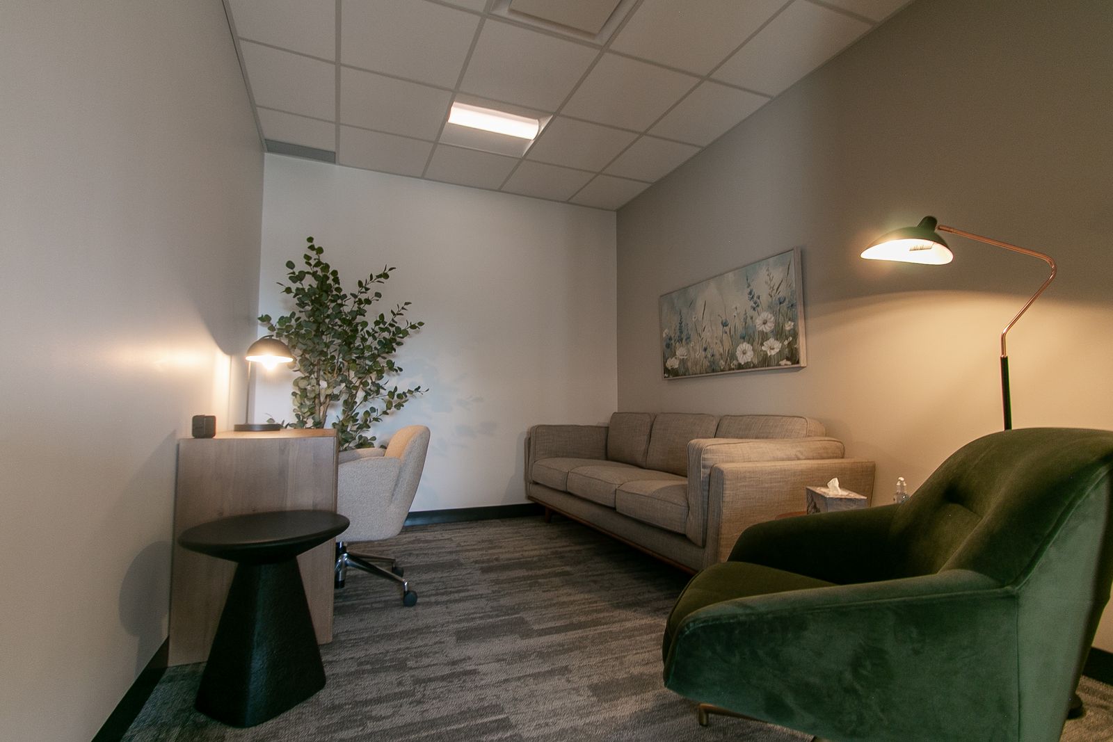 A therapy room at Clinic Studios in lamplight: a sofa, a green velvet armchair, a floor lamp and a desk