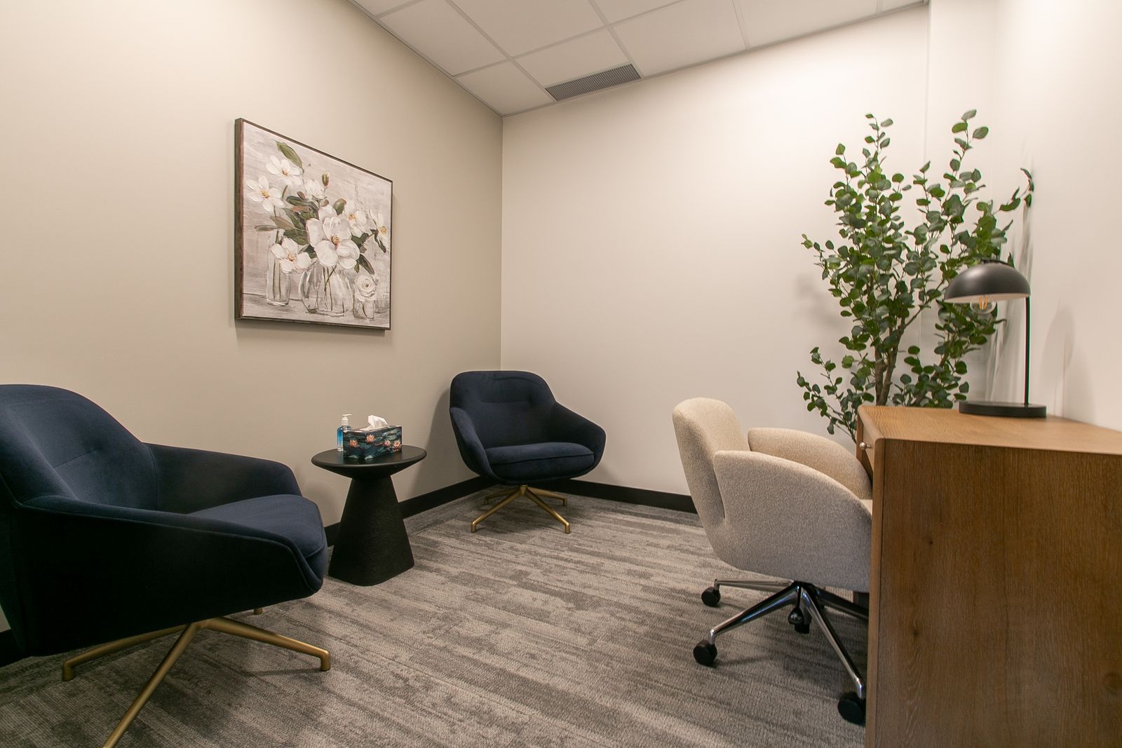 A studio room with two navy armchairs, a desk and chair, a tall plant and framed floral art