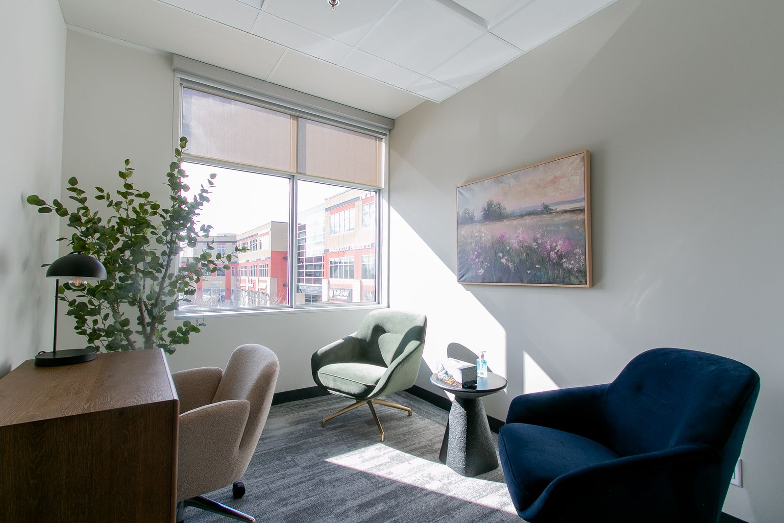 A sunlit studio room with a window, a green chair, a navy chair, a plant and a framed landscape painting